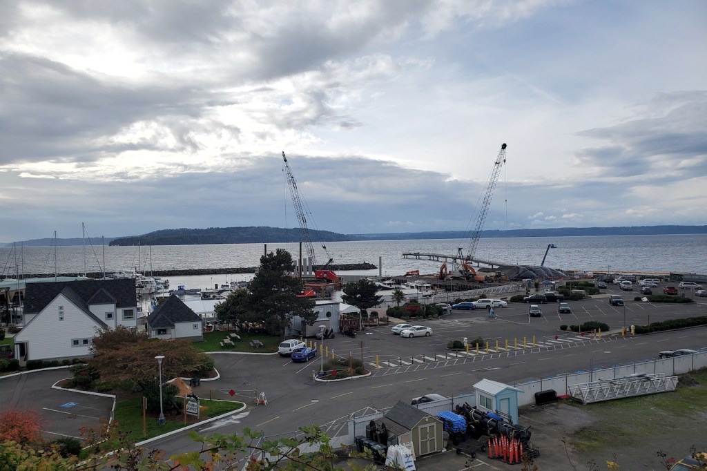 A marina with a parking lot, buildings, two cranes and a pier extending into the water with an island beyond.